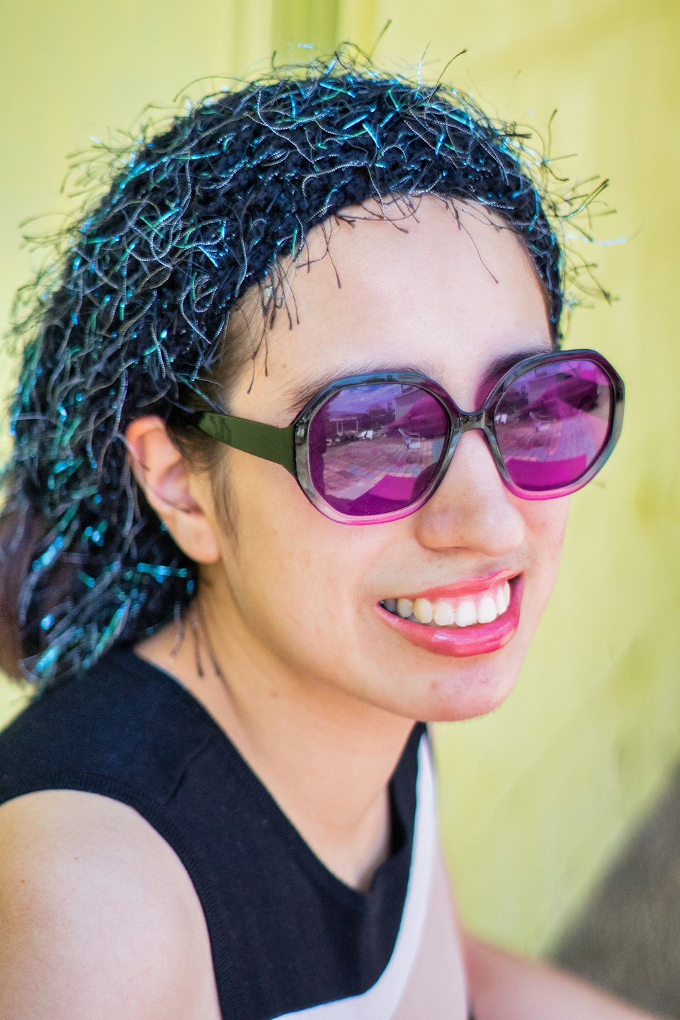 Person wearing purple sunglasses and sparkly head covering smiles against yellow background.
