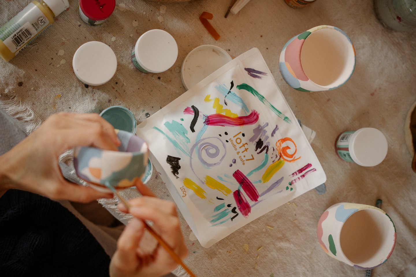 Hands painting abstract designs onto a ceramic cup at a craft table.